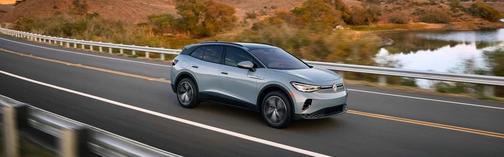The image shows a silver Volkswagen ID.4 electric SUV driving on a scenic road by a lake.