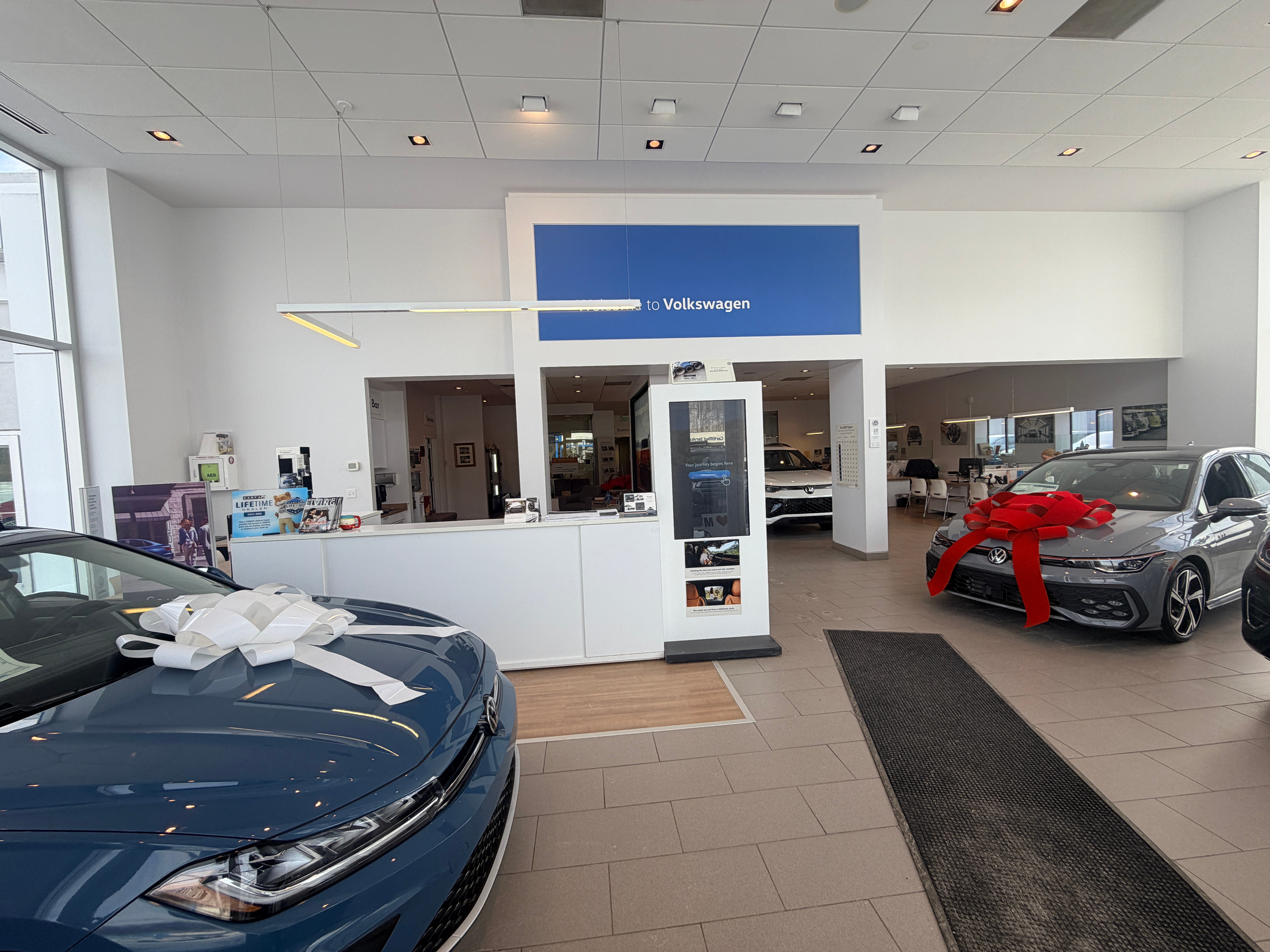 A blue and a gray Volkswagen, both with large bows, on display in a bright showroom.